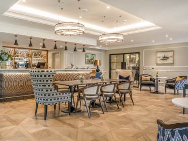 Modern hotel lounge with a bar counter, stylish chairs, a wooden table, and contemporary lighting fixtures. The space is decorated with art, plants, and a neutral color palette.